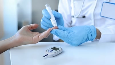 Doctor with gloves measuring blood glucose by piercing patient's finger with lancet and glucometer placed on table